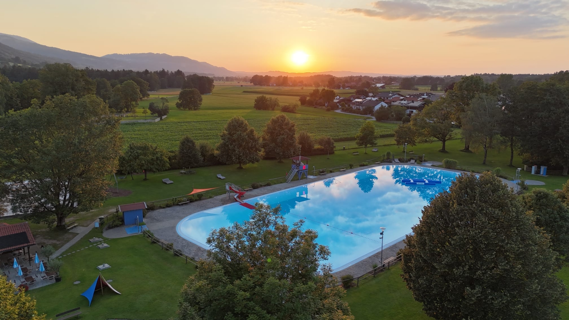 Luftaufnahme des Freibads in Raubling bei Sonnenuntergang mit Schwimmbecken, Liegewiese und Blick auf Felder und Berge.