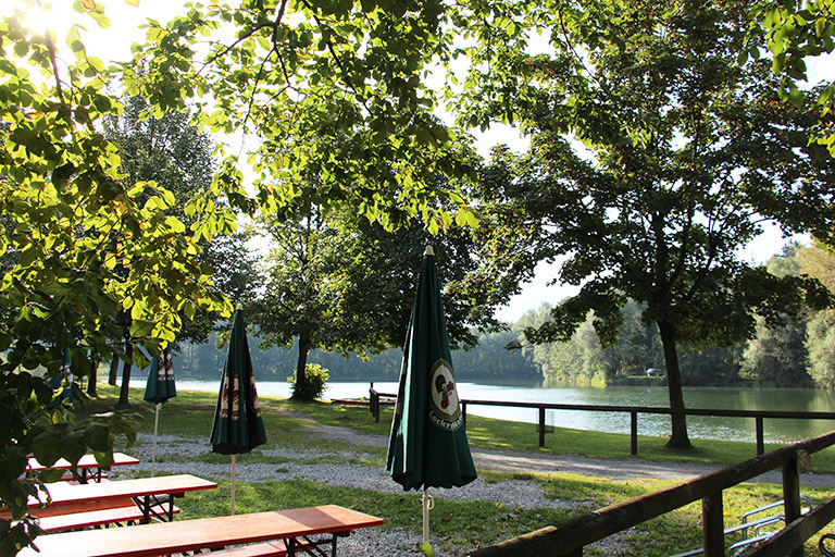 Biergarten mit Tischen und Sonnenschirmen am Ufer eines Sees, umgeben von Bäumen und Grünflächen.
