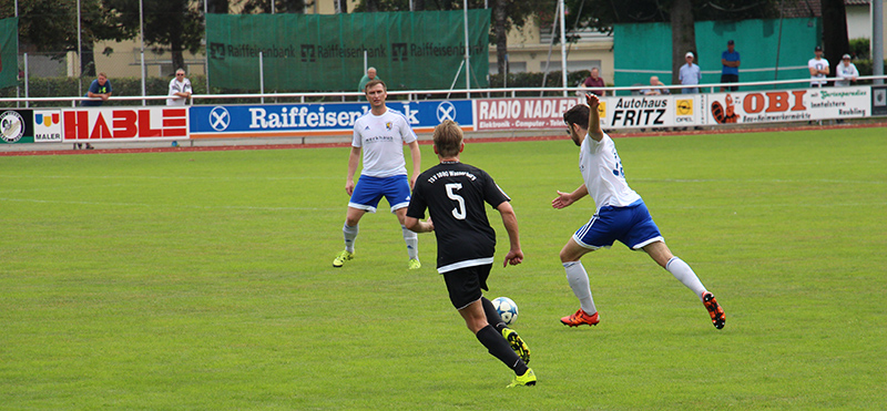 Fußballspiel im Stadion: Drei Spieler kämpfen auf dem Rasen um den Ball, Zuschauer und Werbebanden im Hintergrund.