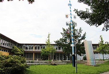 Schulgebäude des Gymnasiums in Raubling mit Innenhof, Grünfläche und Maibaum im Vordergrund.