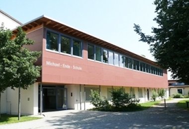 Schulgebäude mit roter Fassade und großen Fenstern, Eingang mit Beschriftung „Michael-Ende-Schule“.