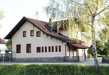 Schulgebäude mit hellem Putz und Satteldach, umgeben von Bäumen und Grünfläche.