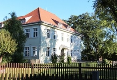 Schulgebäude mit heller Fassade und rotem Walmdach, umgeben von Garten und Holzzaun.