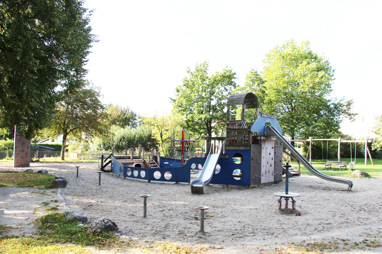 Spielplatz mit Klettergerüst und Rutsche im Sandbereich, umgeben von Bäumen und Grünflächen.
