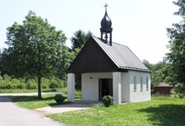 Kleine Kapelle mit dunklem Dachreiter und weißer Fassade, gelegen an einer Wiese mit Bäumen.