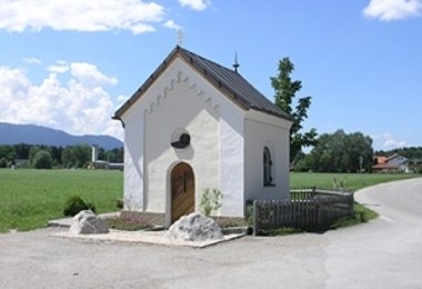 Kleine Kapelle mit Satteldach und Holztür umgeben von Wiese und Bergen im Hintergrund.