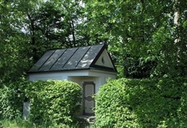 Kleine Kapelle mit dunklem Dach, umgeben von dichtem Grün und Hecken.