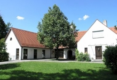 Gebäudekomplex mit rotem Ziegeldach und Innenhof mit Rasenfläche und Baum, umgeben von Hecken.