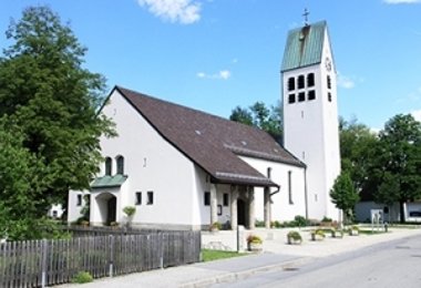 Kirche mit weißer Fassade und hohem Turm an einer Straße, umgeben von Bäumen.