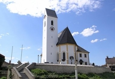 Kirche mit weißem Turm und rundem Chor auf einer Anhöhe, mit Treppe und Mauer im Vordergrund.