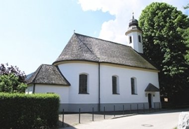 Kleine weiße Kirche umgeben von Hecke und Bäumen.