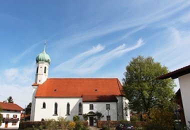 Kirche mit rotem Satteldach und grünem Zwiebelturm, umgeben von Wohnhäusern und Bäumen.