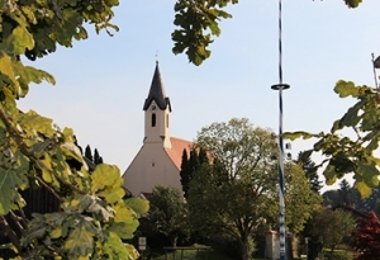 Kirche mit schlankem Turm und spitzem Dach, umgeben von Bäumen.