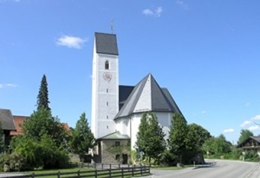 Kirche mit weißem Turm und steilem Dach an einer Dorfstraße, umgeben von Bäumen und Wohnhäusern.